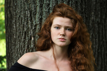 Portrait of ginger girl with long red hair and freckles standing near the tree in summer park. Young sensual model, natural female beauty