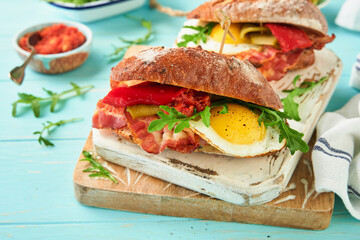 Sandwich layered. English or American freshly prepared picnic sandwich with egg, arugula, bacon, pickles and tomato sause on b turquoise table background on sunny day, copy space