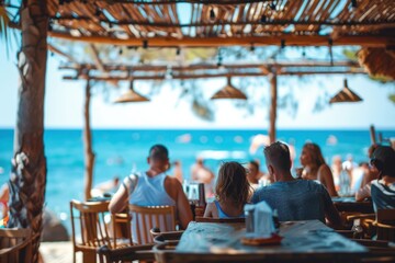 A family sits in a beachside restaurant under a bamboo canopy, enjoying food and drinks while gazing at the ocean, with other patrons and beachgoers in the background.