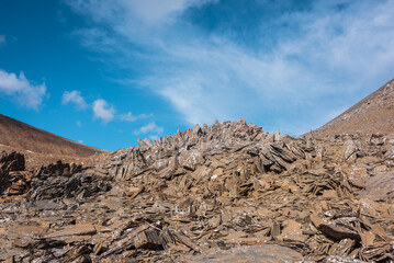 Sunlit stone hill in high mountain valley under clouds in blue sky. Pile of stones on mountain pass in bright sun. Rocky hill of round shape in sunny day. Stony mountain top in changeable weather.