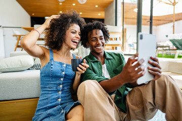 A young couple of African descent is sitting in a modern living room, both smiling and enjoying...