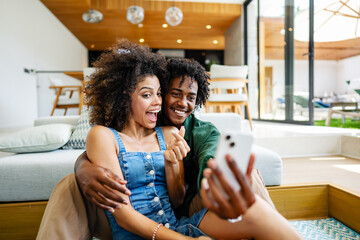 A young couple of African descent is sitting in a modern living room, both smiling and enjoying...