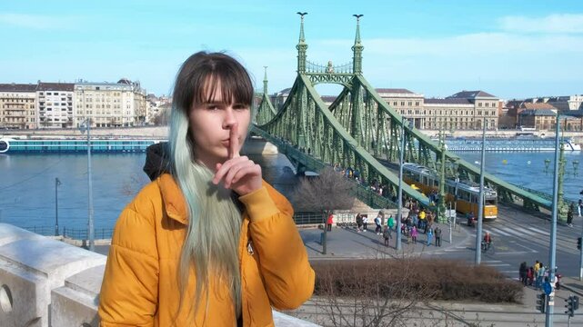 Girl with secret in europe. A view of teen keep secret with her gesture in the street against Danube river.