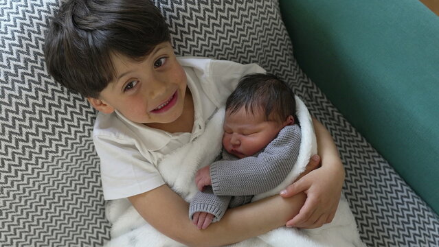 Young boy with a joyful expression holds his newborn sibling wrapped in a cozy blanket, showcasing a moment of pure happiness and brotherly love