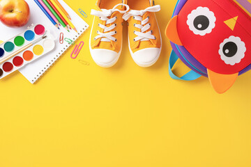 Top view of back to school supplies including shoes, owl backpack, notebooks, apple, and paints on a bright yellow background