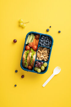 Top vertical view of a healthy school snack in a lunchbox with fruits, vegetables, and nuts on a yellow background