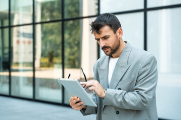 Fototapeta premium Young business man team leader manager standing in front of office building with digital tablet. Portrait of successful designer professional outside work place in financial district. Businesspeople