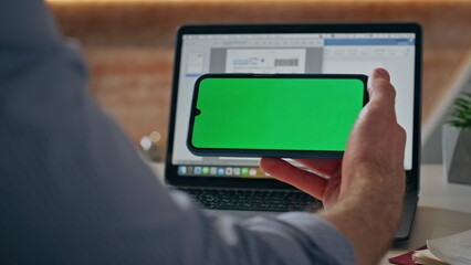 Man finger tapping green screen smartphone at office laptop work desk closeup
