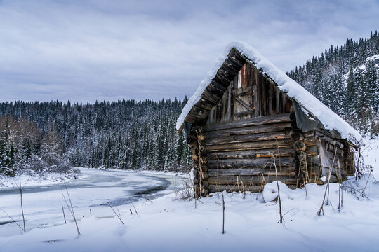 The old wooden cabin deep in the woods at Usva, Perm krai of Russia, with the snowy forest (taiga) and frozen blue river.
