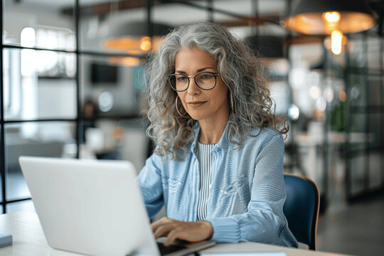 Mature female entrepreneur working on laptop in modern office, older businesswoman with grey curly hair and glasses typing on computer at desk. Professional woman managing digital project