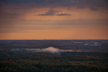 A serene sunrise over a forested landscape with mist hovering above the trees. The sky glows with soft orange hues, creating a peaceful and picturesque scene.