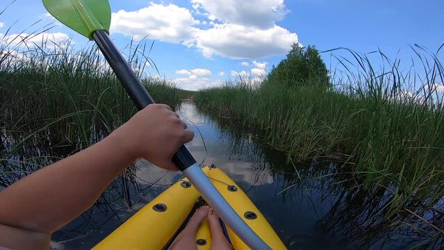 A woman floating in an inflatable boat on a river through green thickets reeds on a summer day. One person rowing oars in a boat. POV. Kayaking on the calm river. Tourism, recreation, entertainment