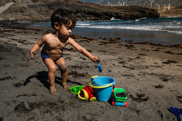 A young boy is playing in the sand on a beach. He is holding a bucket and he is digging in the sand. The beach is near a body of water, and there are several wind turbines in the background