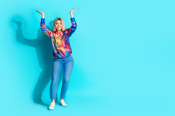 Full size photo of lovely girl dressed print shirt raising hands up presenting object empty space isolated on blue color background