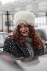 beautiful long-haired brunette in furry white hat and black coat is smiling sweetly on street