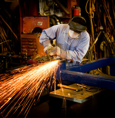 A man creating bright sparks while grinding metal in a machine shop. The man is wearing a long sleeve blue shirt and blue jeans