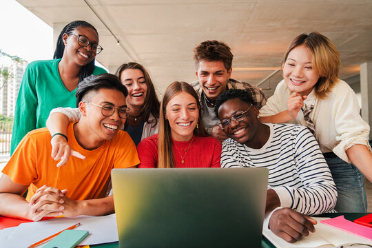 Big group of young students using a laptop to do the homeworks at high school library. Teenagers e-learning with a computer at university campus. Classmates talking and studying for a exam at class