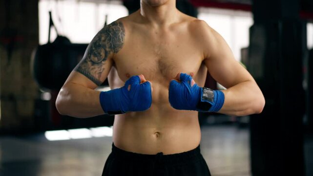 close-up in boxing fight club, a bald young boxer with tattoos in blue bandages stretches his hands before a fight professional training