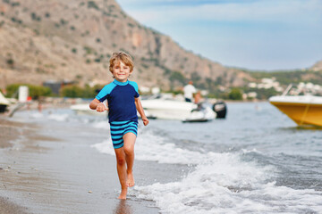 Adorable little blond kid boy running on ocean beach. Child having fun near sea. Vacations, summer, travel concept. Summer family vacations on Mediterranean Sea