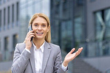 Confident businesswoman in gray suit talking on mobile phone outside modern office building. Cheerful and professional woman having business conversation outdoors.