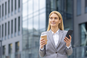 Fototapeta premium Confident businesswoman standing outside modern office building holding coffee cup and smartphone. Professional attire and focused expression depict success, business communication