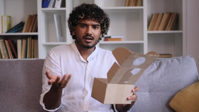 Handsome man received parcel dissatisfied with shopping in online internet store at home Upset guy customer disappointed looking at camera with purchase order while open carton box indoors