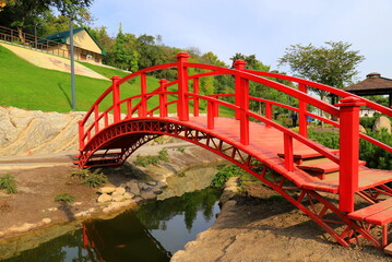 Old red wooden bridge across small river in park. Vintage Japanese style bridge, water. Uman park, Ukraine