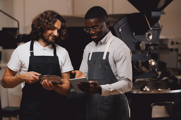 Team work of professional roasters. Afro american man use computer tablet, caucasian barista hold coffee beans after roasting machine, food factory.