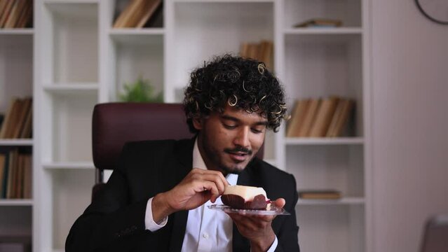 Cunning young indian businessman looking around to make sure nobody watching and eating cake with great pleasure at home Smiling male biting and enjoying bakery indoors Break the diet concept