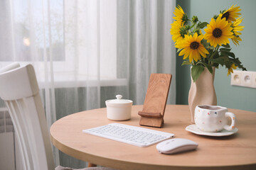  Home workplace in interior - stand for mobile, keyboard, computer mouse, cup of coffee, vaze with sunflowers on wooden table near the window in living room.
