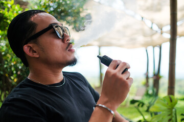 Asian man vaping with an electronic cigarette vape device concept. Smoking with an E-cigarette blowing smoke from mouth, and enjoying outdoors in summer, modern alternative to tobacco addictive habit.