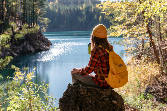 young woman tourist with yellow backpack in checkered shirt sitting on stone on the shore of lake drinking from thermal mug Autumn landscape