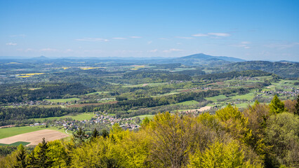 Panoramic view of Jested-Kozakov Ridge from Kozakov Mountain in Bohemian Paradise, Czechia, showcasing a lush green landscape with trees and rolling hills under a clear blue sky.