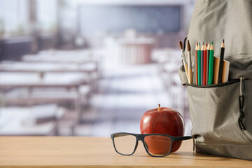 Back to school concept. Wooden desk top with backpack and apple and school stuff with blurred classroom backround 