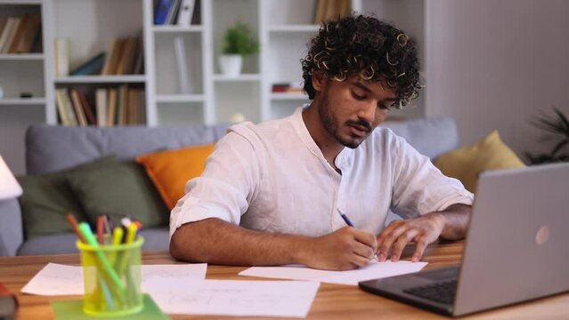 Busy indian guy preparing for exam at university studying at home workplace Positive student make notes while looking at laptop computer listening lesson indoors Distance remote education	