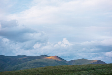 Fototapeta premium Scenic view from grassy flowering hill to high multicolor mountain under cloudy sky. Sunlit mountain top of sunset vivid colors in changeable weather. Dramatic twilight landscape with colorful tops.