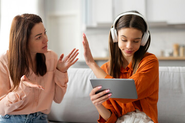 Mother quarreling with daughter, teenage girl in headphones ignoring mom and playing video games on tablet, sitting on sofa at home