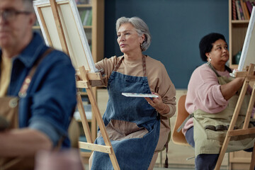 Portrait of elegant senior woman painting on easel in art studio with group of elderly people enjoying creative hobby in retirement home