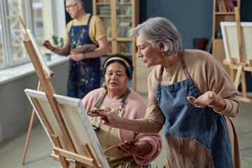 Side view portrait of elegant senior woman giving comments to people painting pictures on easel in art class during creative workshop copy space