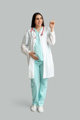 Beautiful young pregnant doctor pointing at something on grey background