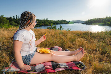 A girl in blue sunglasses sits on the river bank and eats French fries. Sunny summer day in the countryside. Outdoor picnic