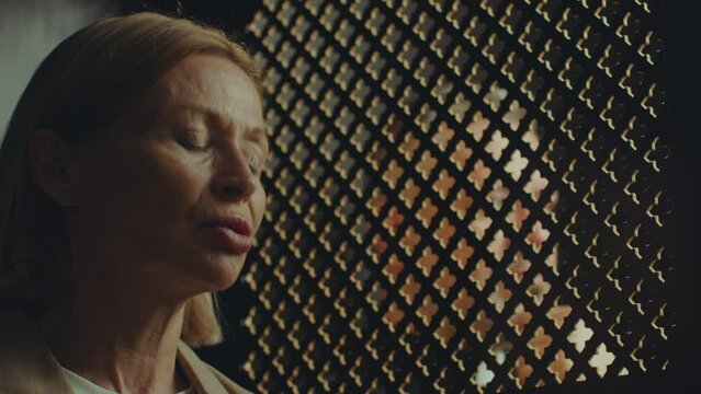 Mature female penitent confessing to priest sitting in confessional booth and listening to her through latticed window