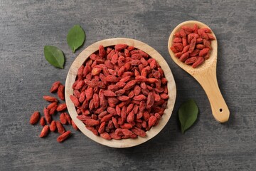 Dried goji berries in bowl, spoon and leaves on grey textured table, flat lay