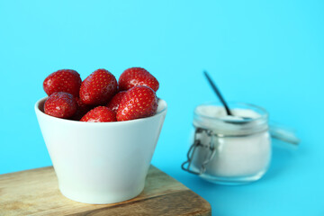 Bowl with fresh sweet strawberries and jar of sugar on blue background