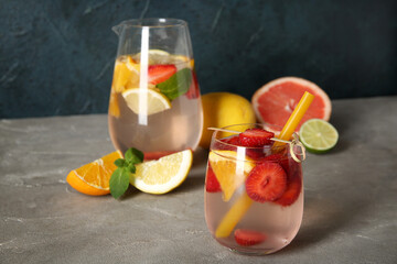 Glass and jug of fresh infused water with strawberries and different citrus fruits on grey table