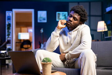 BIPOC bored man passing time at home by watching TV show on laptop. Tired person feeling apathetic in neon lit apartment at night while consuming uninteresting media content