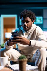 Man texting internet friends using mobile phone in warm stylish home, having fun. Joyful person relaxing in apartment by scrolling on smartphone, entranced by social media activity