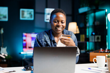 Excited black woman seated at desk doing online shopping on laptop and using debit card for cashless payment. African american lady holding credit card, spending money, doing e commerce banking.