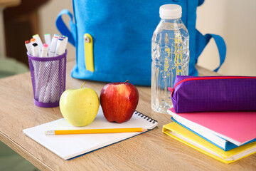 Apples with school stationery, water bottle and backpack on desk in classroom, closeup