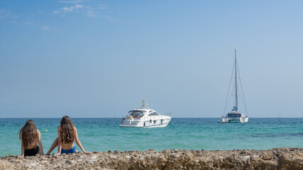 Two young girls, relax on the rocky beach, two ships in the background, azure water